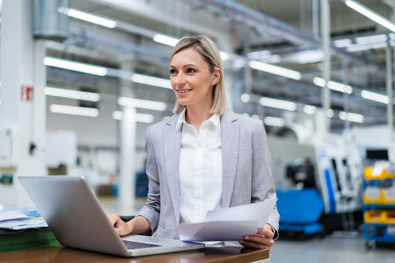 Smiling businesswoman with laptop and documents in factory