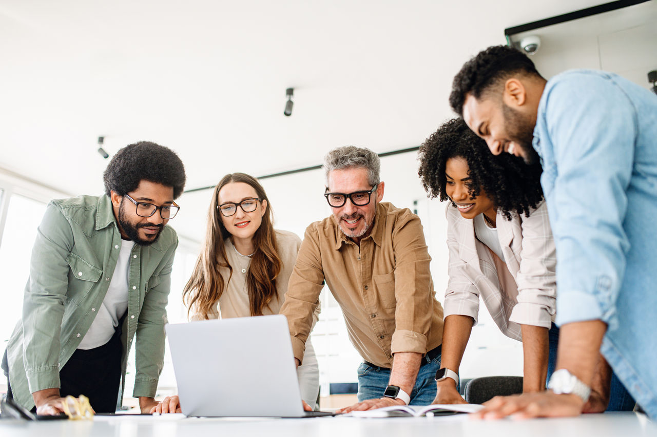 A group of diverse professionals is standing around a laptop planning digital strategy