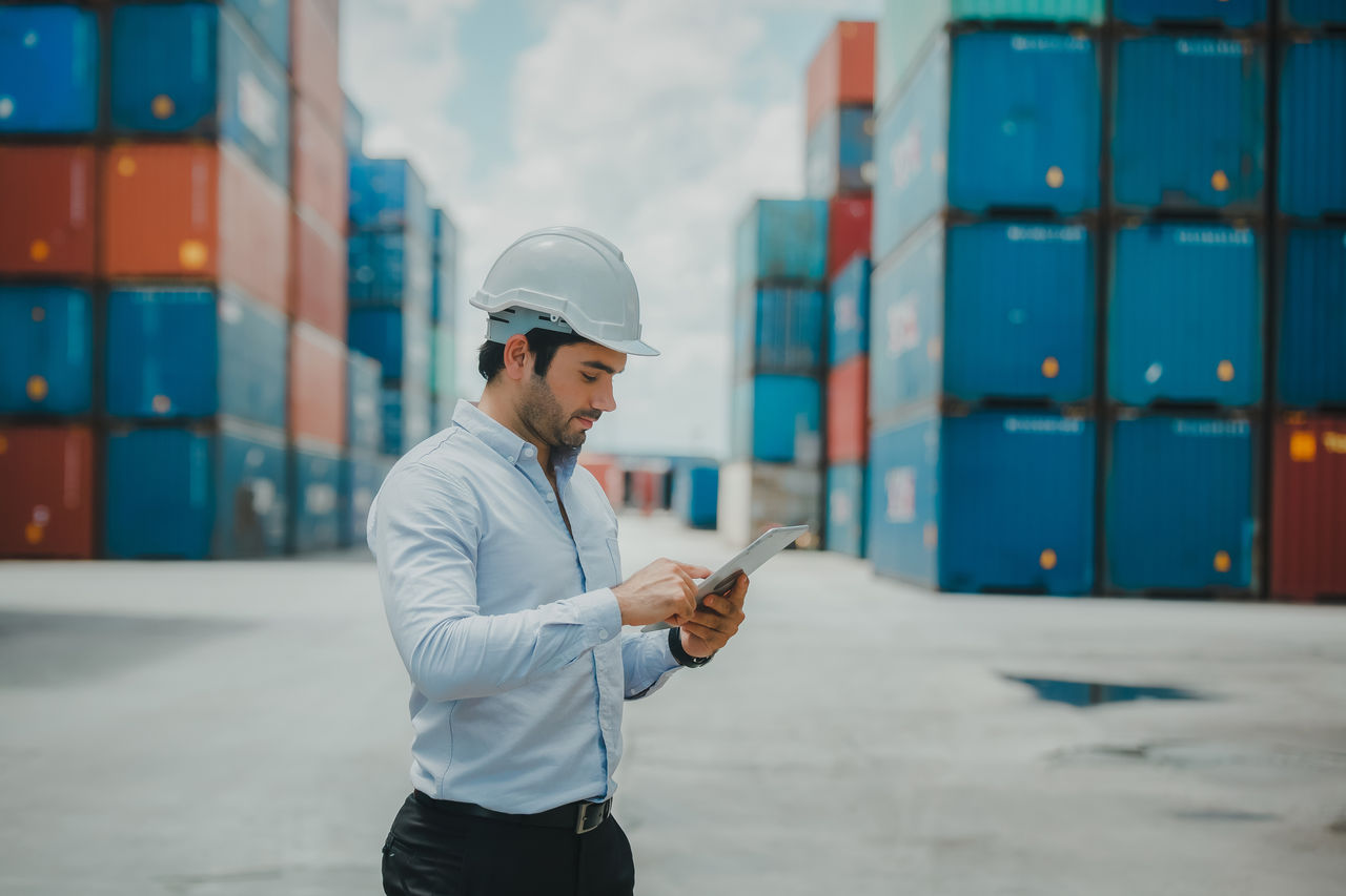 Engineer worker in causual suit standing in shipping container yard holding laptop with smile. Import and export product. Manufacturing transportation and global business concept